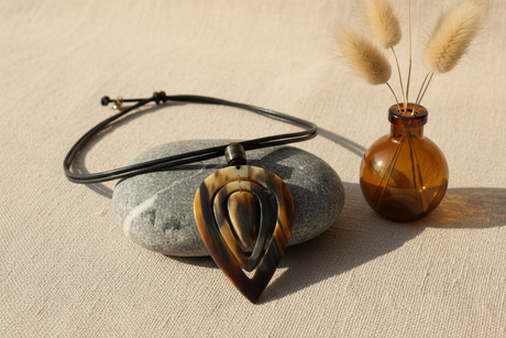 Haitian horn pendant necklace draped over river stone beside pampas grass and amber bottle on linen surface